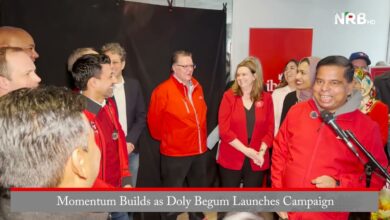 Group of people in red jackets smiling during a campaign kickoff, with a speaker at a microphone in front of a banner that reads Momentum Builds as Doly Begum Launches Campaign.