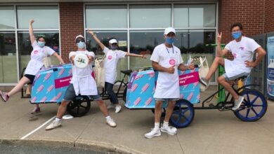 Six people in white aprons and masks pose with a pink popsicle-themed pedicab outside a brick storefront, some arms raised in celebration.