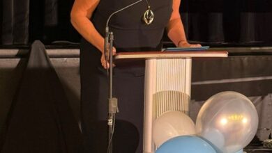 Woman in a black dress speaks at a lectern on a stage, with blue and white balloons nearby.