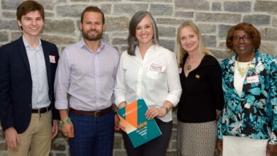 Five adults—three women and two men—smile at the camera while posing in front of a stone wall; the center woman holds teal and orange pamphlets as event materials.