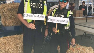 Two police officers in reflective vests pose inside a tent, holding signs with event hashtags/handles (#SupercrawlHFN, @HamOntMarket).