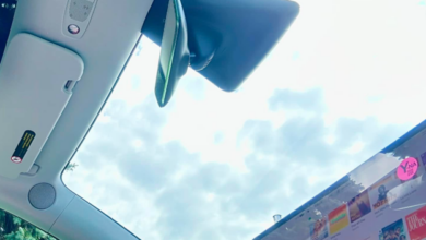 Inside a car cockpit: large touchscreen on the dashboard shows app icons while the steering wheel and rearview mirror are visible against a blue sky outside the windshield.