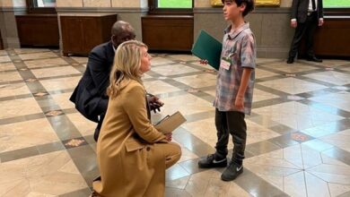 A blonde woman kneels to speak with a young boy holding a green folder in a formal hallway with large portraits on the walls, a man crouched nearby.