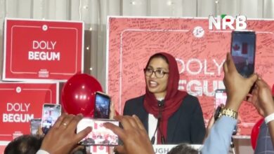 Woman in a red hijab and glasses speaks at a podium during a campaign event with a red backdrop reading 'Doly Begum' and NRB logo; audience records with smartphones nearby.