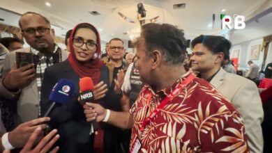 Woman in a red hijab being interviewed by reporters, microphones in front of her at a crowded indoor event.