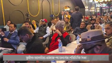 Crowded indoor event with attendees seated and listening, some recording with smartphones; a man in a red vest and knit cap sits near center, foreground/right a man wearing a hat; Bengali news ticker at the bottom and NRB HD watermark on screen.