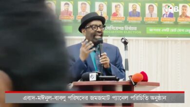 Man in a blue suit and teal shirt speaks into a microphone at a podium during a political press conference, with several microphones in front and a banner of candidate photos in green behind him.