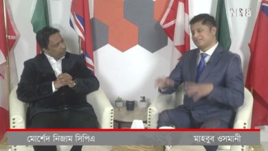 Two men in suits sit in cream chairs on a studio set, with international flags in the background and a Bengali caption bar across the bottom.