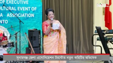 A woman in a peach sari speaks into a microphone on a stage with musical instruments and a blue backdrop behind her, NRB HD logo in the corner, and Bangla caption ticker at the bottom