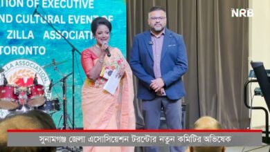 Female host in a peach sari speaks into a microphone on stage, with a man in a blue blazer beside her; drums and a blue banner are behind them.
