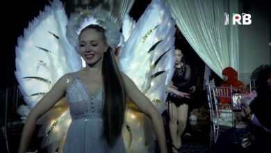 Woman in a white gown with large illuminated feather wings walks down a backstage runway, smiling as onlookers film with a smartphone nearby.