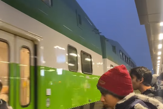 People boarding a green and white train at a station platform, with a yellow tactile strip along the edge and passengers in winter coats and a red knit hat.
