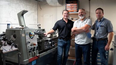 Three men in a machine shop standing beside a large gray metal lathe, with one man pointing at controls and smiling.