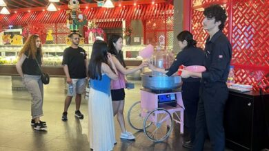 Group of young customers queue for cotton candy in a bright dessert shop with red lattice walls and decorative hanging lights, pink cotton candy cart in use by staff.