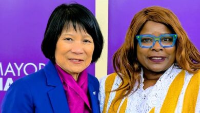 Two women pose for a photo in front of purple campaign banners; one in a royal blue suit and pink scarf, the other in a white lace top with a yellow scarf and turquoise glasses.