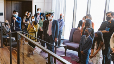 Group of people wearing masks standing in a bright modern lobby with large windows, watching a presentation.