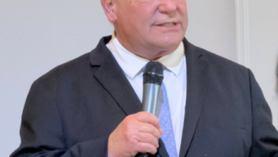 Man in a dark suit and light blue tie speaks into a handheld microphone at a formal event.