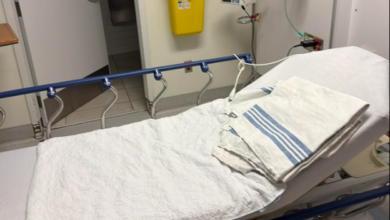 Hospital bed with white sheets and a folded striped towel, in a patient room with IV equipment and a yellow sharps container on the wall.