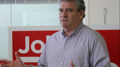 Man in a light purple button-down shirt speaks with open hands, standing in front of a red campaign sign behind him.