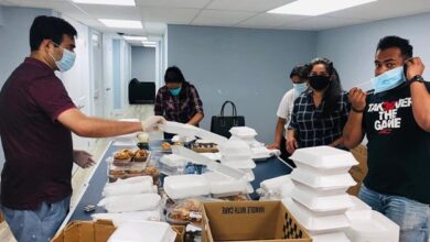 Volunteers wearing masks in a blue-walled room package meals on a table with stacks of white takeout boxes and supplies.