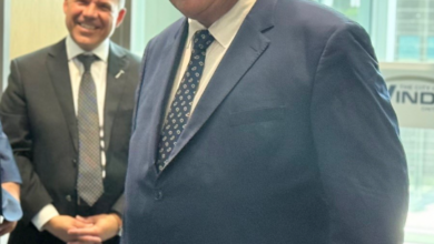 Smiling man in a navy suit with a patterned tie stands in an office by a window, with another suited man in the background.