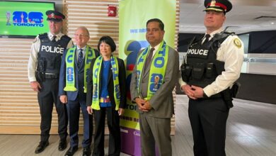 Group of five people posing for a photo in a lobby: two uniformed police officers at the ends, three civilians in business attire wearing blue-green scarves, with a bright banner behind them.