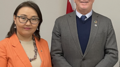 Two professionals pose for a photo indoors, a woman in an orange blazer beside a man in a gray suit with a flag and official emblem behind them.