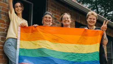 Four friends stand on a brick porch, holding a rainbow Pride flag and smiling at the camera.