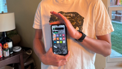 Person in a white T‑shirt holds a smartphone upright against their chest, screen showing app icons on the home screen.