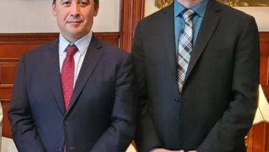 Two men in business suits standing side by side and smiling in a formal room with framed paintings behind them.