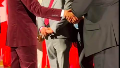 Man in a burgundy suit shakes hands with a man in a gray suit on a stage, red curtains behind them, with a podium and microphone at the front.
