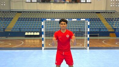Young male athlete in a red sports kit poses with thumbs up in front of a blue futsal goal inside an empty indoor arena.