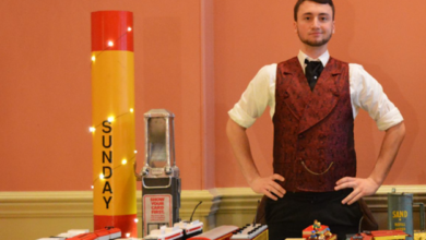 Man in a burgundy vest stands behind a table of toy trains and a tall red-yellow pillar labeled 'SUNDAY' on display.