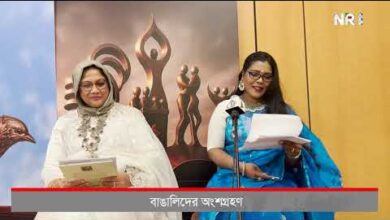 Two women in traditional outfits stand at microphones on a stage, holding papers, with a decorative backdrop and a Bengali caption bar.