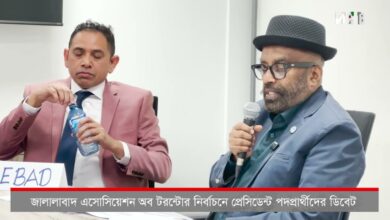 Panel discussion with two men: left, in a pink suit, opens a water bottle; right, in a hat and glasses, speaks into a microphone, Bengali caption bar at bottom.