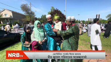 Reporter from NRB TV interviews a group of families in colorful traditional South Asian clothing at an outdoor gathering on a sunny day. A man holds a child while another man holds a microphone toward him, and several women and children stand nearby with houses and a crowd in the background.