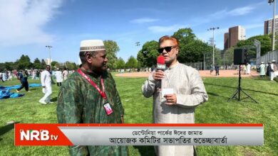 Reporter with NRBtv microphone interviews a man in white traditional attire on a grassy field; a sports ground and other people in background.