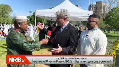 Reporter interviews a suited man with a NRB TV microphone at an outdoor park event; another man in white traditional clothing stands nearby under a tent.