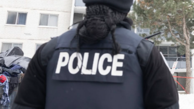 Back view of a police officer wearing a dark vest labeled POLICE in a snowy urban setting with apartment buildings in the background.