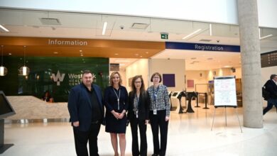 Four professionally dressed people stand together in a bright lobby near Information (orange sign) and Registration (blue sign) desks, with a WestHealth logo on the wall behind them.