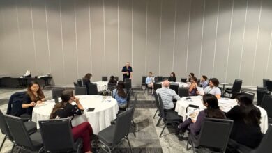 Presenter stands at the front of a conference room while attendees sit around round tables, listening to a speaker.
