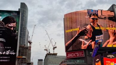 Crowded Times Square street with towering digital billboards, including a racer with the caption 'TOGETHER WE FLY HIGHER' amid cheering pedestrians.