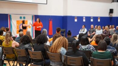 Audience seated in a gym watching a stage presentation; two women on stage with a colorful banner behind them.