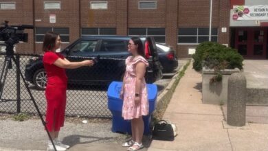 Reporter in a red dress interviews a student in a pink dress outside a brick school building, with a video camera on a tripod nearby.