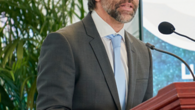 Man in a dark suit and light blue tie delivering a speech at a wooden podium with a microphone, indoors near leafy plants, wearing glasses.