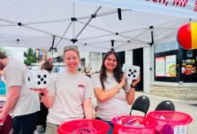 Two smiling volunteers stand under a tent banner reading 'Mark Gerretsen' while holding oversized dice at a street fair booth with red buckets on the table behind them.