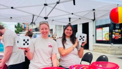 Two smiling volunteers stand under a tent banner reading 'Mark Gerretsen' while holding oversized dice at a street fair booth with red buckets on the table behind them.