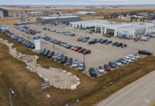 Aerial view of a large car dealership lot with hundreds of cars arranged in long rows, a white showroom building, a Canadian flag on a tall pole, and surrounding development.