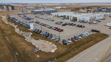 Aerial view of a large car dealership lot with hundreds of cars arranged in long rows, a white showroom building, a Canadian flag on a tall pole, and surrounding development.