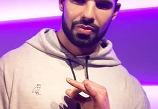 Man with a beard in a light gray hoodie making a hand gesture against purple neon background.